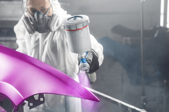 Male automobile repairman painter in protective workwear and respirator painting part of car body to rare purple color in paint chamber