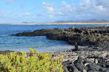 A white man wearing black shorts and a black T-shirt walks on a volcanic beach in the Canary Islands - Fuerteventura. He looks at the waves of the blue Atlantic Ocean