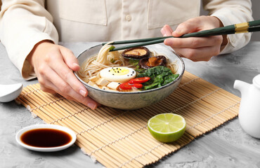 Woman eating delicious ramen with chopsticks at grey table, closeup. Noodle soup