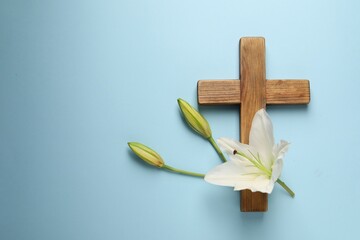 Wooden cross and lily flowers on light blue background, top view with space for text. Easter...