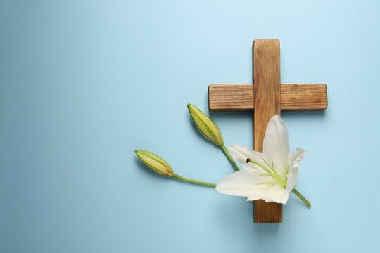 Wooden cross and lily flowers on light blue background, top view with space for text. Easter attributes
