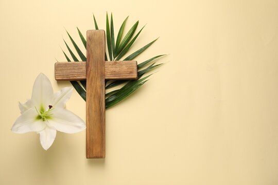 Wooden cross, lily flower and palm leaf on pale yellow background, top view with space for text. Easter attributes - Powered by Adobe