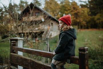 Young girl in warm clothes sitting on aged wooden fence, wooden gate, looking at autumn nature.