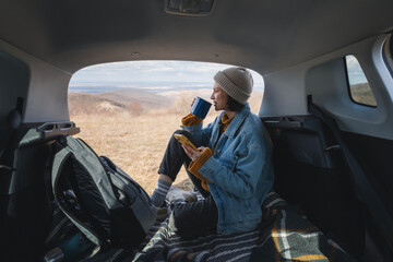 A young happy woman sitting in an open car trunk drinking tea from a mug. Travel by car concept