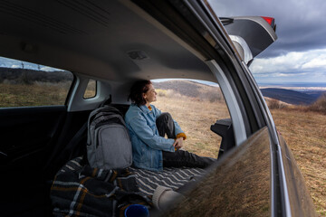 Young cheerful woman traveler sitting in open trunk of car enjoying nature& Solo travel concept - 745647716