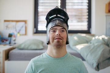 Portrait of smiling man with down syndrome at home with knight's helmet on head.
