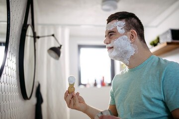 Young man with down syndrome learning how to shave, applying shaving foam all over his face,...