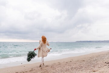 Redhead woman Christmas tree sea. Christmas portrait of a happy redhead woman walking along the beach and holding a Christmas tree in her hands. Dressed in a light coat, white suit and red mittens.