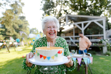 Beautiful senior birthday woman with paper crown holding birthday cake. Garden birthday party for...