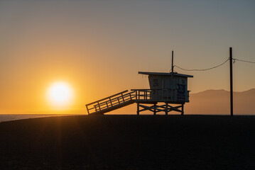 A lifeguard tower stands silhouetted against a colorful sunset on a beach. The sky is ablaze with...