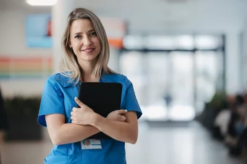  Portrait of confident female doctor in hospital corridor. Beautiful nurse wearing blue uniform, holding clipboard standing in modern private clinic, looking at camera. © Halfpoint