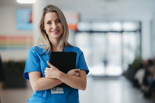 Portrait Of Confident Female Doctor In Hospital Corridor. Beautiful Nurse Wearing Blue Uniform, Holding Clipboard Standing In Modern Private Clinic, Looking At Camera.