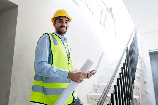 Smiling architect wearing hardhat and standing on steps holding laptop at site