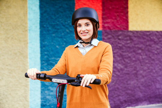 Smiling woman standing with electric push scooter in front of wall