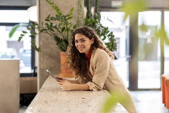 Smiling young businesswoman with tablet PC leaning on cabinet at workplace