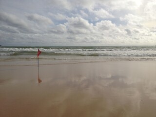 strong waves in the sea and the red flag with no swimming sign on the clear sand beach seeing cloud's reflection on it