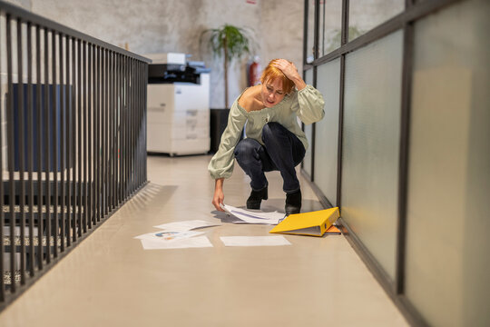 Careless mature businesswoman picking scattered documents in corridor at office