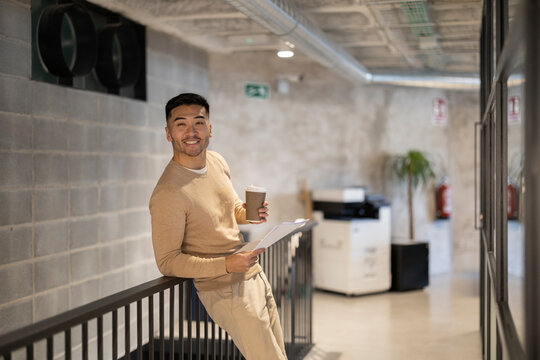 Smiling young businessman with disposable coffee cup and documents leaning on railing at office