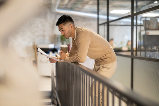 Young businessman with disposable coffee cup and documents leaning on railing at office