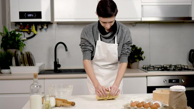 Cute teenage boy vigorously kneads dough with hands making effort in beautiful cozy kitchen. Ingredients flour eggs butter for shortbread cookies with milk on table. Cook boy kneads dough giving it