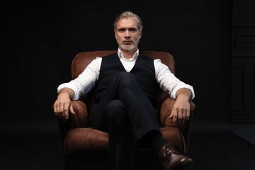 In leather chair sitting senior businessman with gray beard wearing dark suit. Against black wall