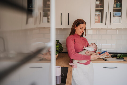 Woman carrying sleeping baby daughter and using smart phone in kitchen