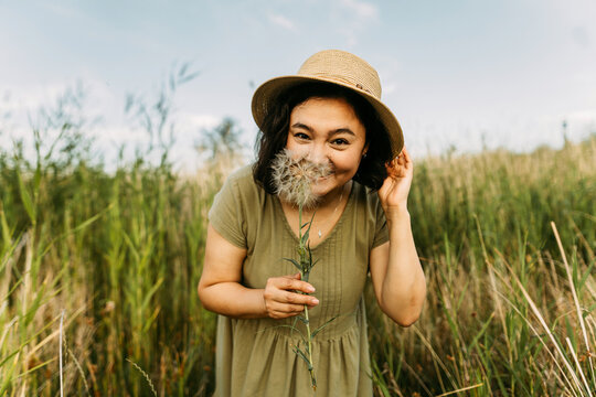 Playful woman wearing hat and holding dandelion near face in field