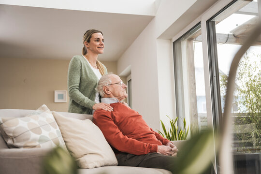 Happy home caregiver with senior man sitting on sofa in living room