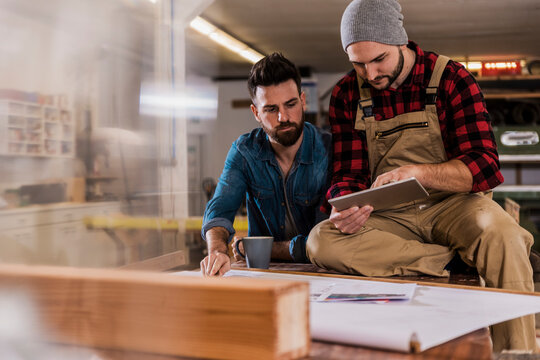 Carpenter sharing tablet PC with colleague sitting on workbench at workshop - Powered by Adobe