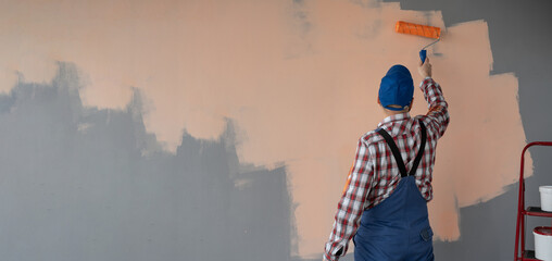 Back view of painter man painting the wall in orange colour, with paint roller, on big empty space. Banner.