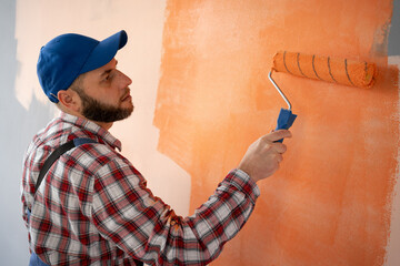 Back view of handsome painter in blue dungarees painting a wall with paint roller in orange color.