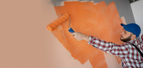 Painter in blue cap and overalls painting a wall with paint roller. Builder worker painting surface with orange color.