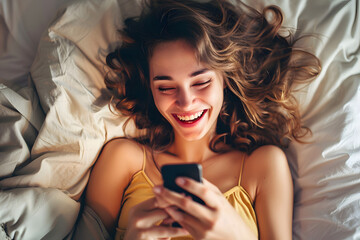 A beautiful young woman lying in bed laughs looking at her smartphone