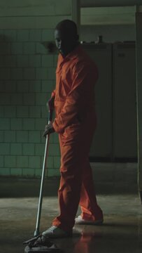 Vertical shot of Black prison inmate in orange jail uniform mopping floor in dark room