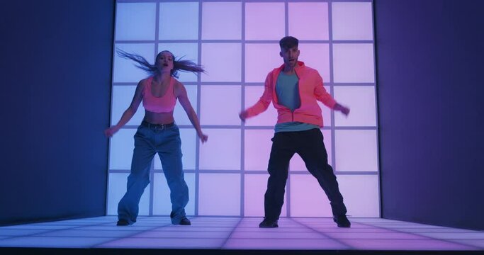 Young cool energetic couple of dancers in casual trendy wear urban hip-hop performing street style choreography on animated luminous neon display radiant neon panels background.