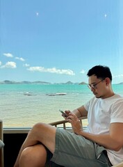 Asian man sitting and using mobile phone near the window with beautiful sea view, working and relaxing alone in cafe on chilling day time.