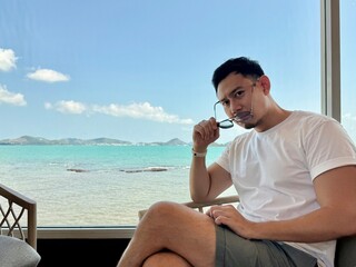 Asian man sitting on chair near the window with beautiful sea view, holding sunglasses while look at camera,relaxing and chilling on holidays, happy life concept.