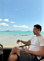 Asian man sitting on chair near the window with beautiful sea view, relaxing and chilling on holidays, happy life concept.