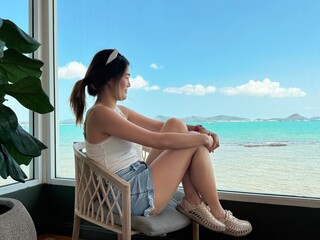 Asian Thai woman sitting on chair near the window with beautiful sea view, relaxing and chilling on holidays, happy life concept.