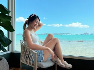 Asian Thai woman sitting on chair near the window with beautiful sea view, relaxing and chilling on holidays, happy life concept.
