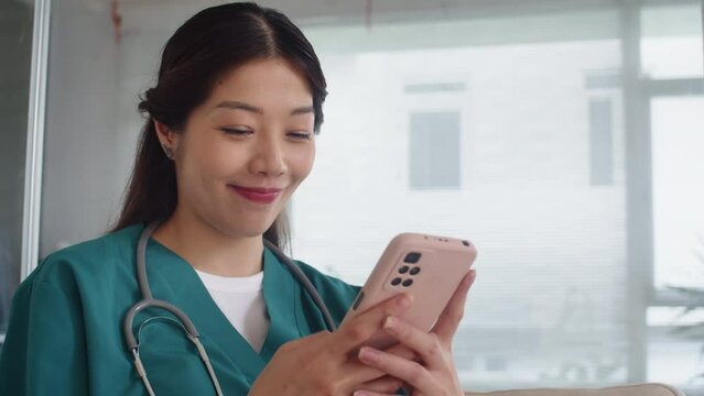Tilt up of happy Asian nurse in scrubs chatting with friends on smartphone while having break at work