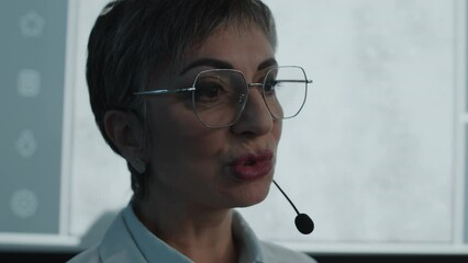 Experienced female doctor in white coat and glasses talking in mic in front of projection screen while giving presentation on healthcare conference