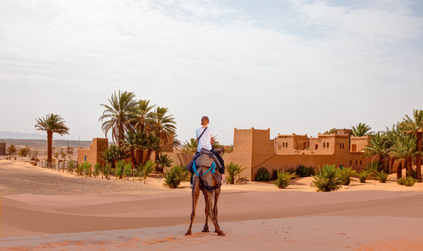 A woman riding a camel across the thin sand dunes of the in Western Sahara Desert - General view of the Merzouga hotels district - Morocco, Africa