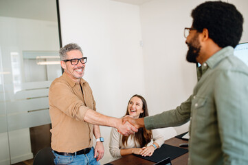 A man in a beige shirt beams as he shakes hands with a colleague, signifying a friendly and...