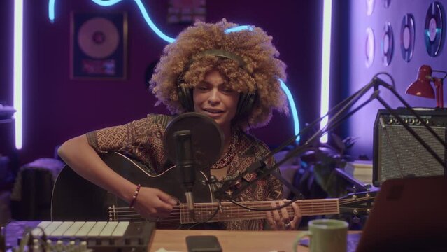 UGC footage of young Black woman with lush hair recording song while singing to guitar at neon lit home studio at night and looking at camera
