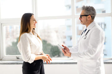 A seasoned doctor with grey hair engaging in a friendly conversation with a young female patient by a window, reflecting a healthcare setting that is both personal and infused with daylight, side view