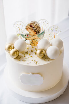 Trendy baptism cake with figure angel with wings and golden decor. Cake on background white photo zone. Celebration baptism concept. Delicious reception at birthday baby party. Closeup. Side view.