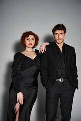 good looking couple in elegant black attires posing together on gray backdrop and looking at camera