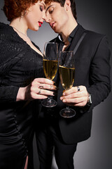 alluring woman in black dress hugging her handsome boyfriend on gray backdrop, champagne in hands