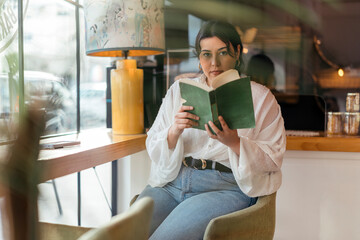Chica joven guapa en cafetería sentada junto a una ventana con un libro 
